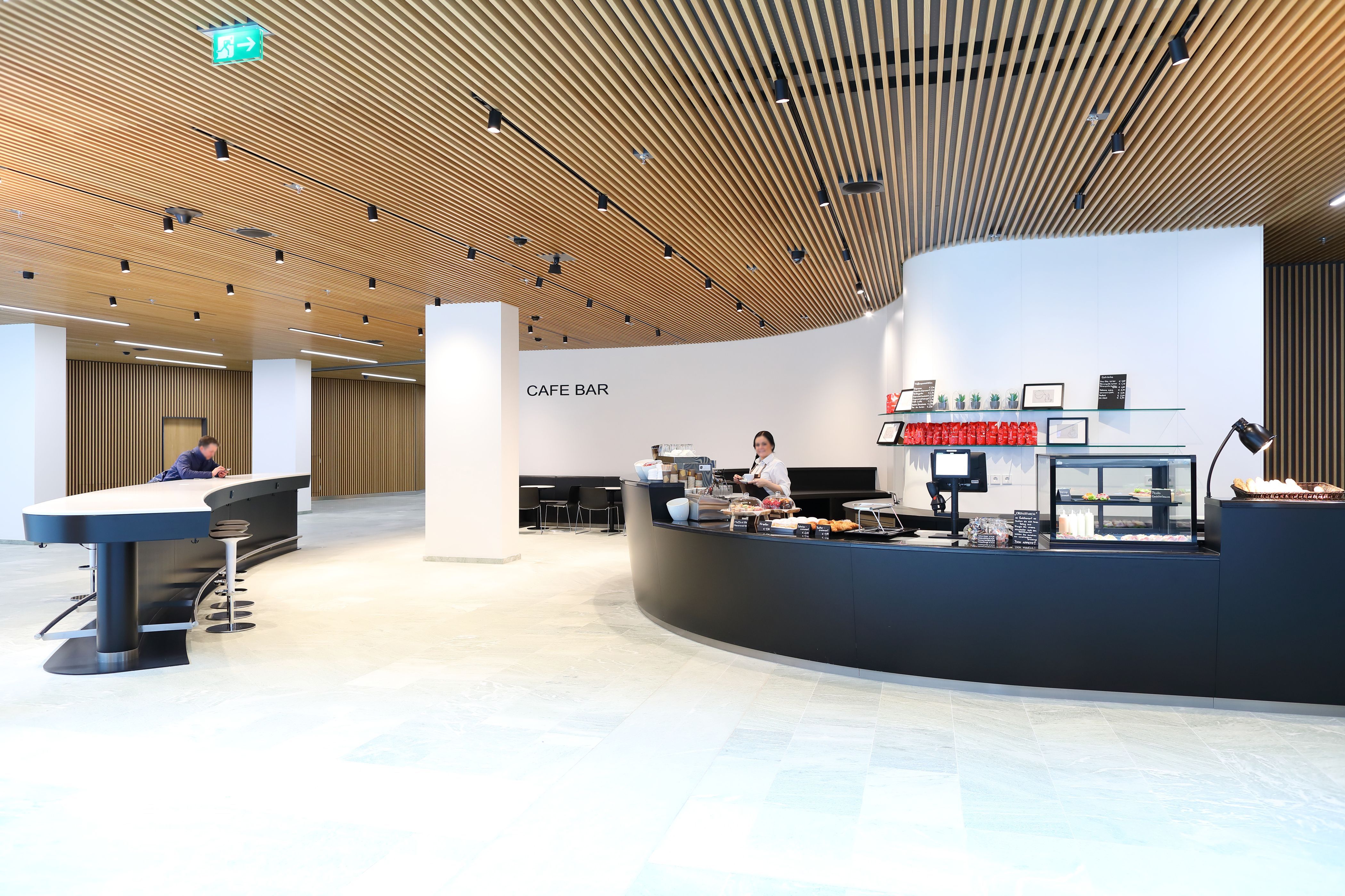 Photo: EURO PLAZA; Restaurant Plaza Eurest; coffee bar, very modern and generously designed, featuring a high curved table; an employee is preparing coffee; a guest with a mobile phone at the table. 