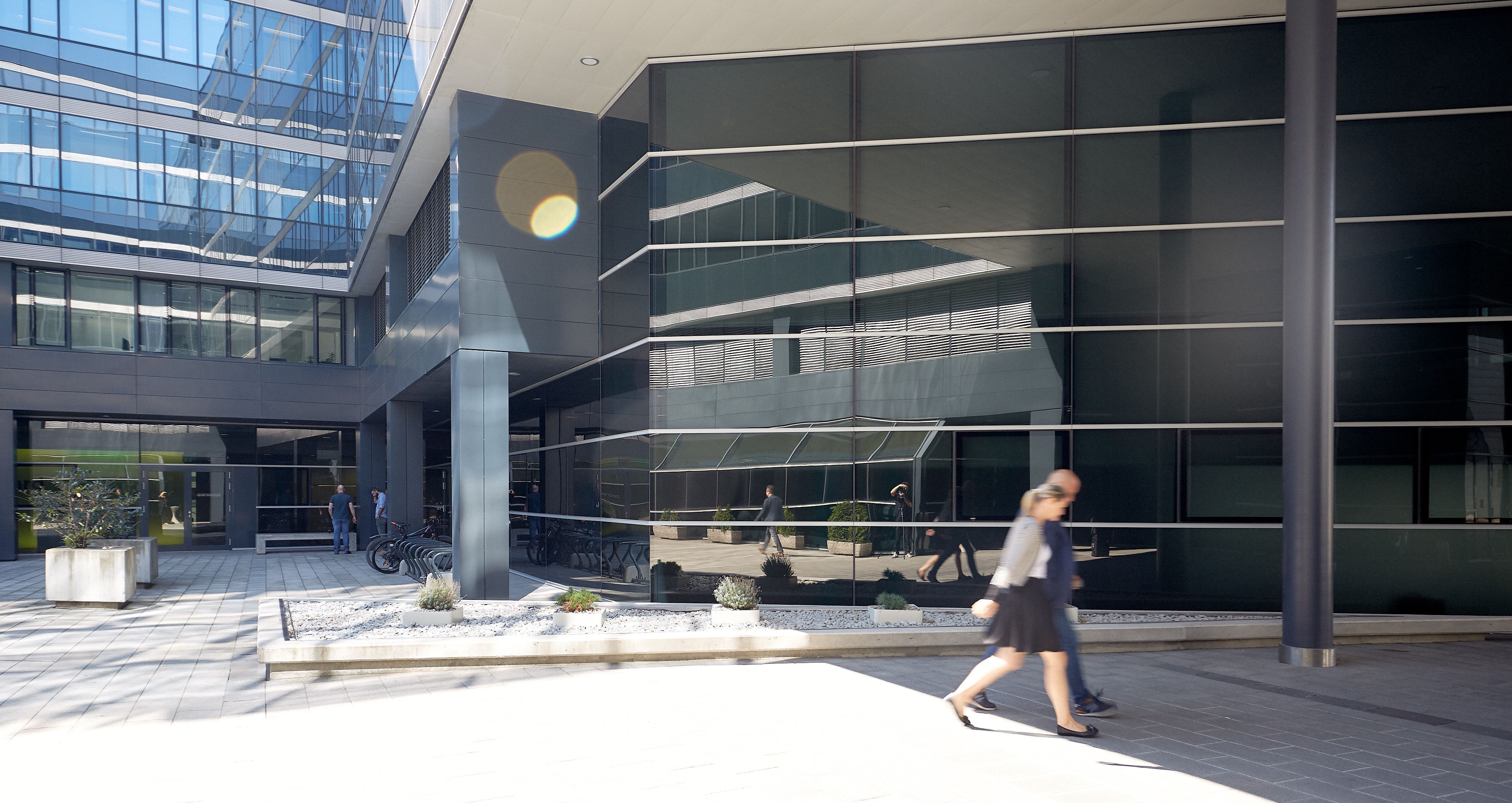 Foto: Ausschnitt eines modernen Bürogebäudes, in dessen Glasfassade sich die Alu-Glasfassade eines anderen Bürogebäudes spiegelt; im Vordergrund eine Frau und ein Mann, die nach rechts gehen 