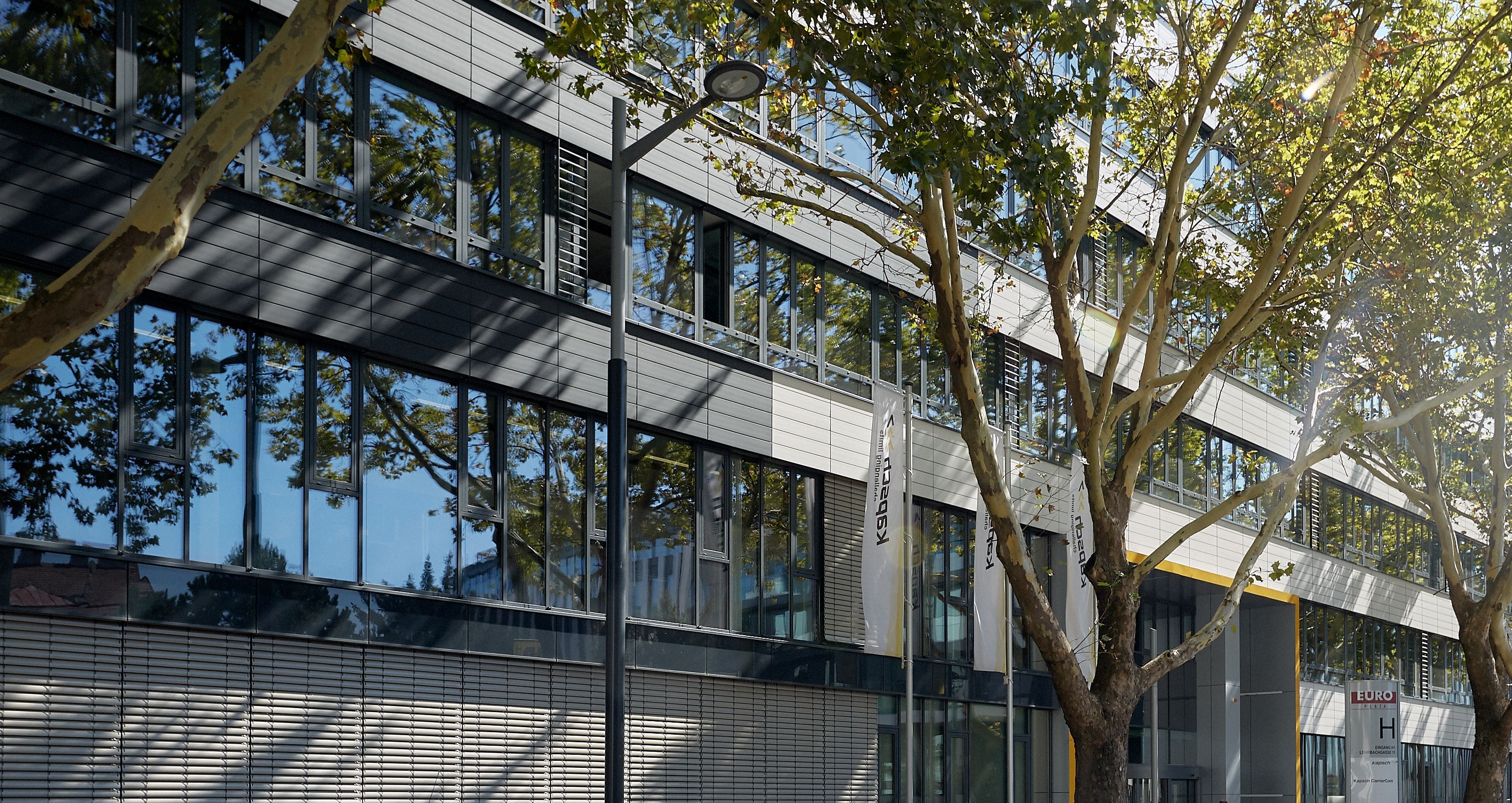 Foto: durch Bäume beschattete Alu-Glasfassade eines modernen Bürogebäudes; vor dem Eingang drei Fahnenmasten der Firma Kapsch und ein Pylon mit dem Buchstaben „H“; im Vordergrund parkende Autos und Motorräder 