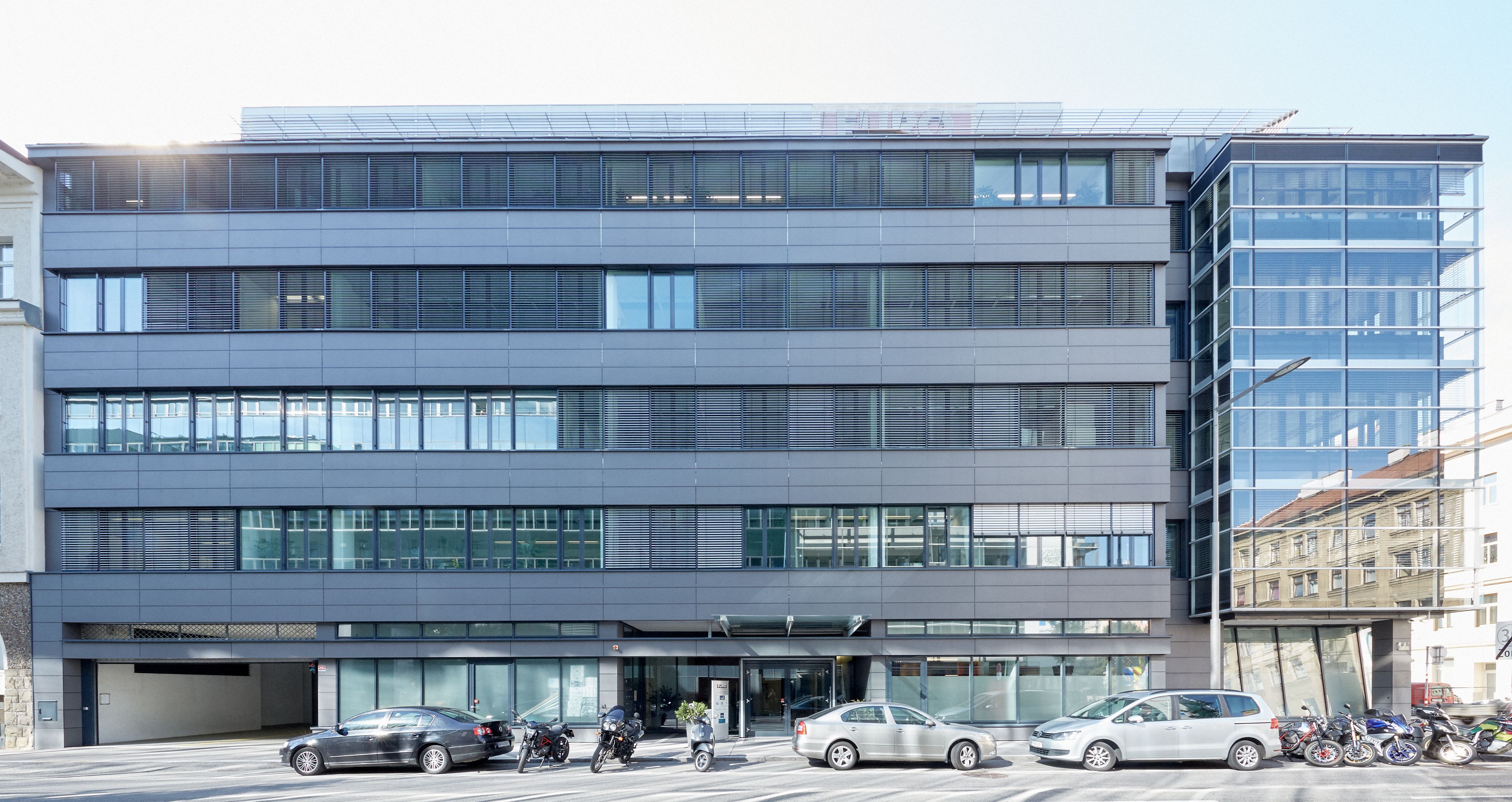 Foto: modernes Bürogebäude mit fünf Geschoßen bestehend aus zwei Teilen: einer mit Alu-Glas-Fassade, der andere mit einer vorgesetzten Glasfront; im Vordergrund parkende Autos und Motorräder