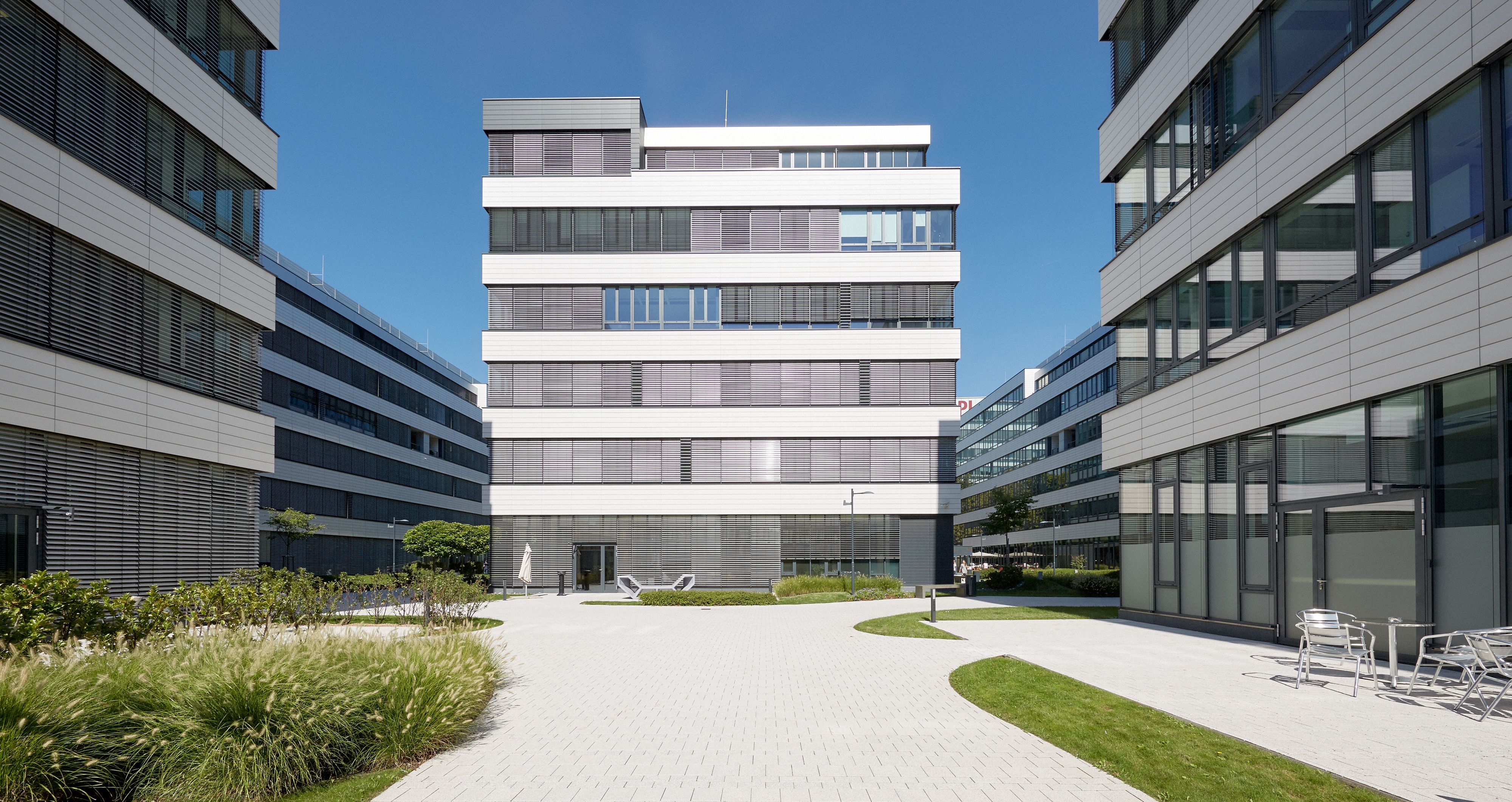 Foto: sauber gepflasterte Fläche umsäumt von Grünflächen und umgeben von hochmodernen Bürogebäuden mit jeweils sechs Geschoßen und Alu-Glasfassade 
