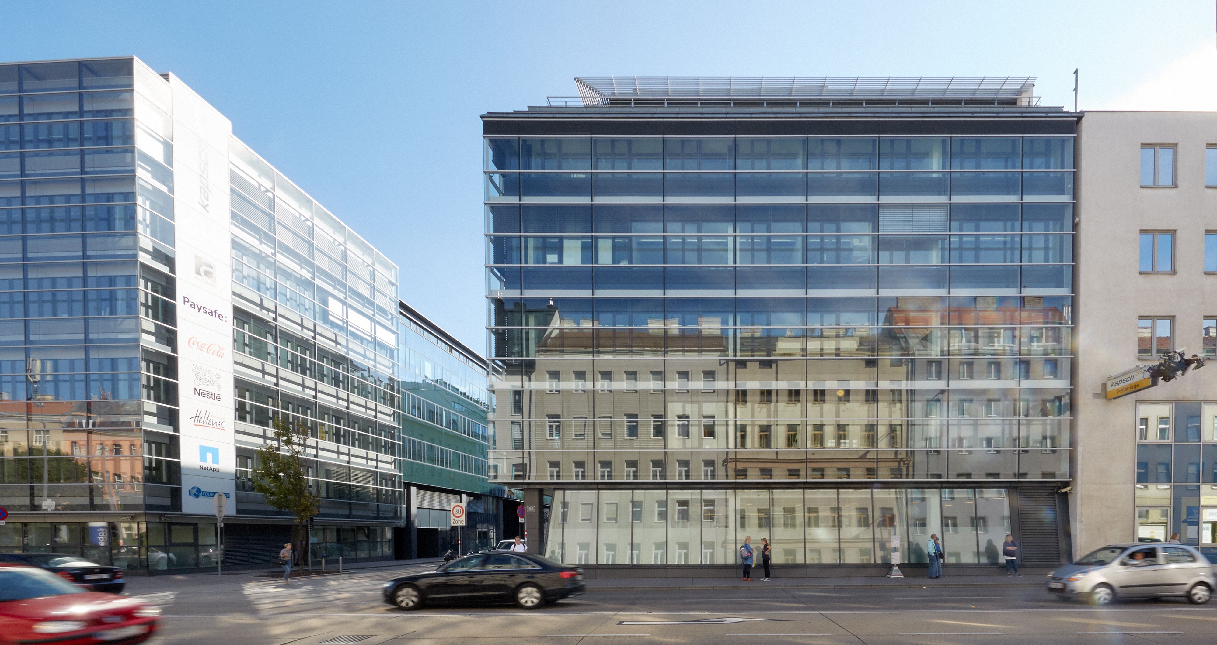 Foto: modernes Bürogebäude mit Glasfassade vor einer stark befahrenen mehrspurigen Straße; in der Glasfassade spiegeln sich bunte Gründerzeithäuser