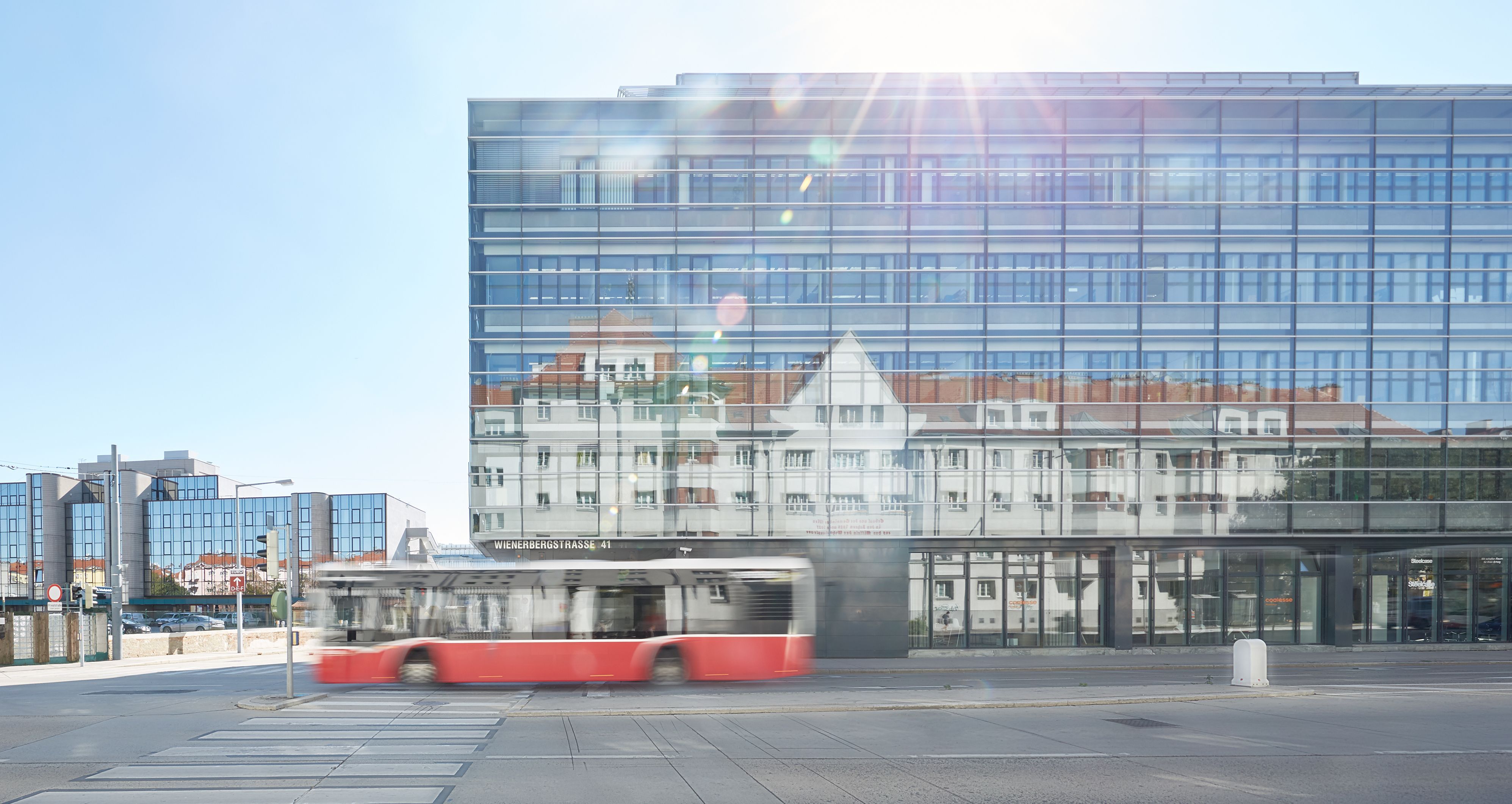 Foto: modernes Bürogebäude mit Glasfassade, in der sich ein gegenüberliegender Altbau spiegelt; davor ein öffentlicher Autobus