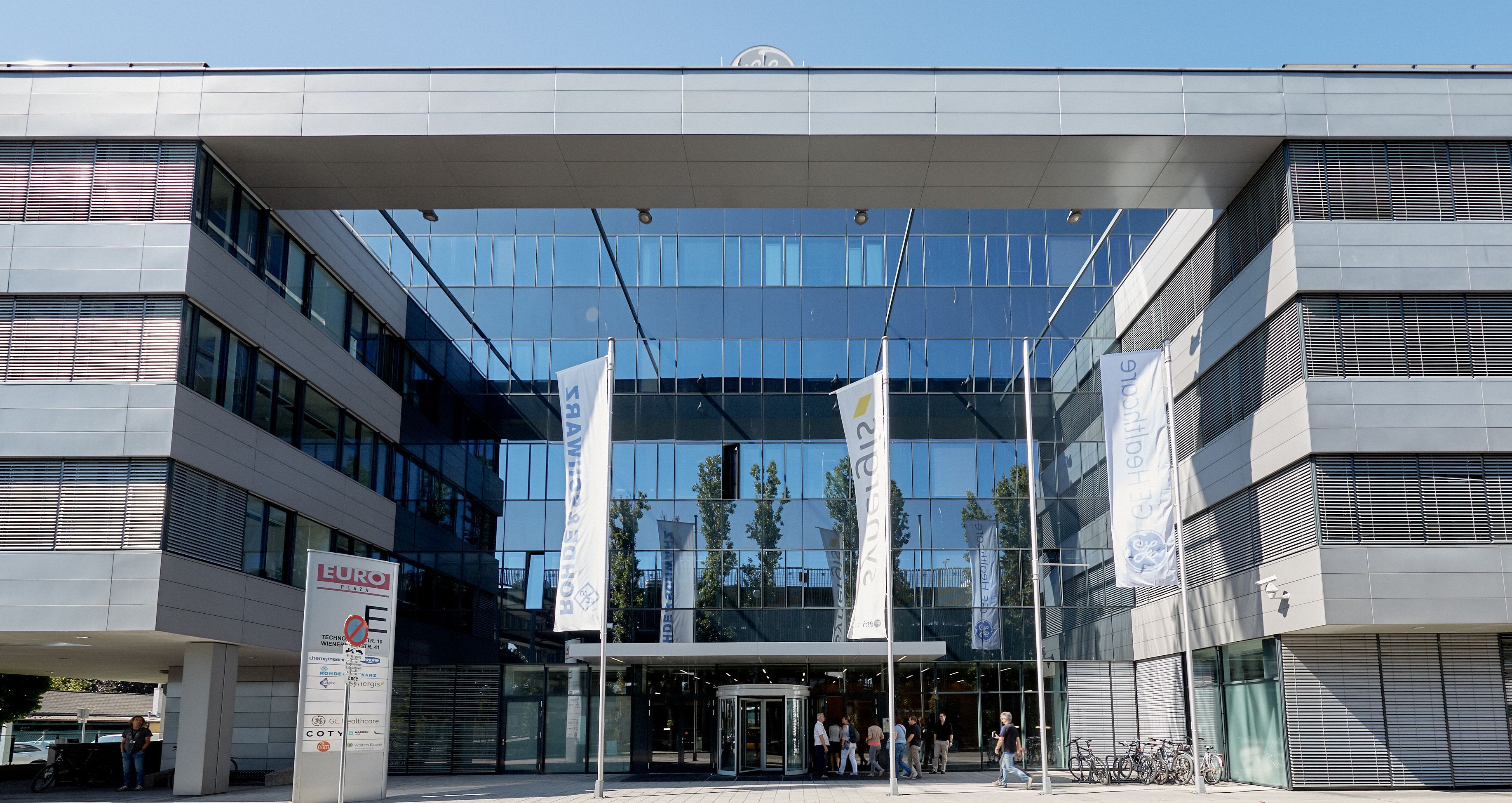 Foto: Eingangsportal eines modernen Bürogebäudes mit Alu-Glasfassade; vor dem Eingangsportal mit Drehtür stehen vier Fahnenmasten, an dreien von ihnen wehen Unternehmensfahnen