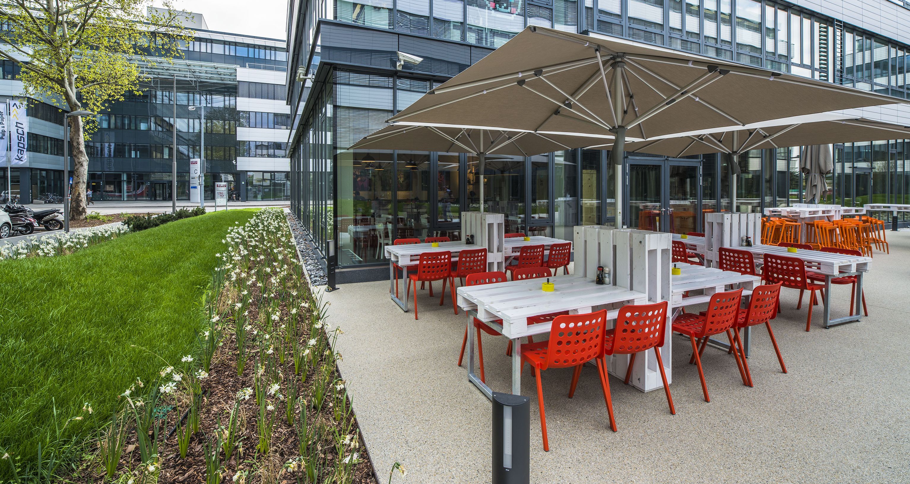 Foto: EURO PLAZA; Restaurant Tuk Tuk; Terrasse neben einer Grünfläche überspannt von einem großen Sonnenschirm; darauf weiße Tische gefertigt aus Europaletten und Metall; rote Stühle.