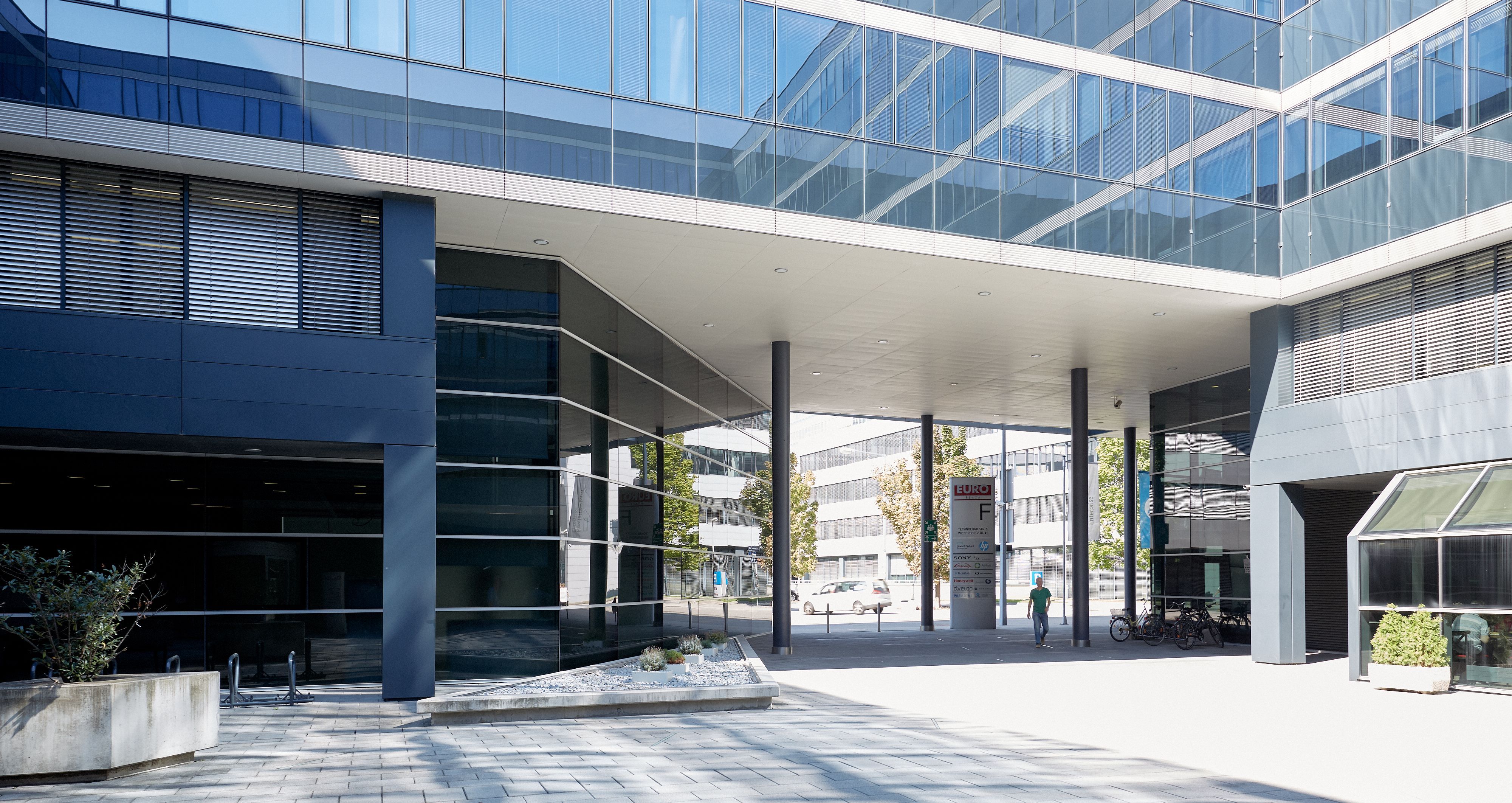 Foto: Durchgang unter einem modernen Bürogebäude mit Alu-Glasfassade; im Durchgang ein Pylon mit dem Buchstaben „F“